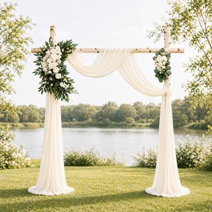 Puede incluir: Un arco de boda de madera decorado con flores blancas, vegetación y tela marfil fluida. El arco está montado sobre un césped con un lago y árboles al fondo, creando un escenario escénico para bodas al aire libre.