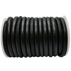 Pode incluir: Um rolo de cordão de couro preto com uma borda costurada. O cordão tem aproximadamente 5 mm de largura e está enrolado em torno de um carretel branco.