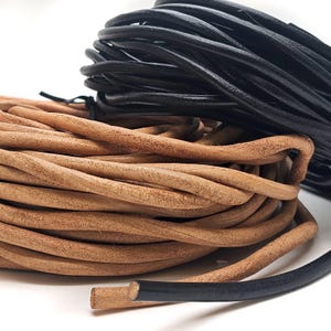 Könnte beinhalten: Zwei Spulen mit Lederkordel, eine braun und eine schwarz. Die braune Kordel ist dicker und hat eine raue Textur. Die schwarze Kordel ist dünner und hat eine glatte Textur.