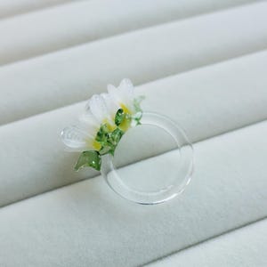Könnte beinhalten: Ein klarer, transparenter Ring mit floralem Design. Der Ring hat eine Ansammlung weißer Blumen mit gelben Zentren und grünen Blättern. Der Ring ist vor einem weißen, strukturierten Hintergrund platziert.