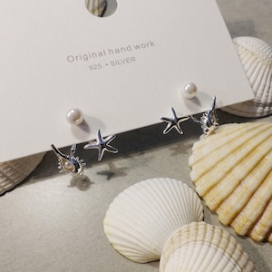 Könnte beinhalten: Ein Set aus drei Paar Silberohrringen auf einer weißen Karte mit dem Text "Original hand work 925 SILVER". Die Ohrringe umfassen Perlenohrstecker, Seesterne und Muscheldesigns. Muscheln umgeben die Karte.