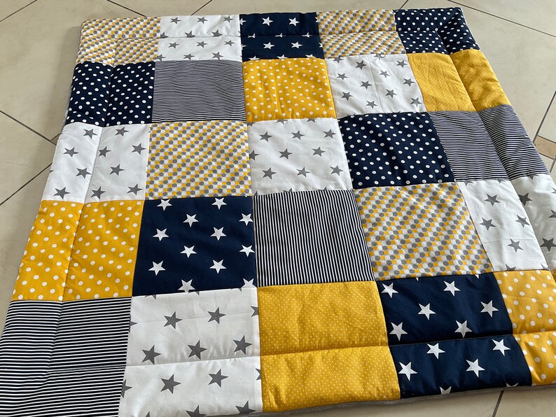 May include: A patchwork quilt featuring a range of patterns in yellow, white and navy blue. The quilt is made up of squares with different designs, including stars, polka dots and stripes.