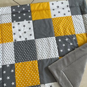 May include: A patchwork baby blanket in a grey and yellow colour scheme. The blanket is made from different fabrics with various patterns, including stars, polka dots and small stars. The blanket has a grey fleece backing.