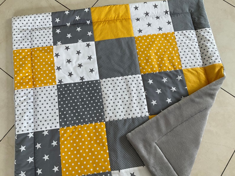 May include: A patchwork baby blanket in a grey and yellow colour scheme. The blanket is made from different fabrics with various patterns, including stars, polka dots and small stars. The blanket has a grey fleece backing.