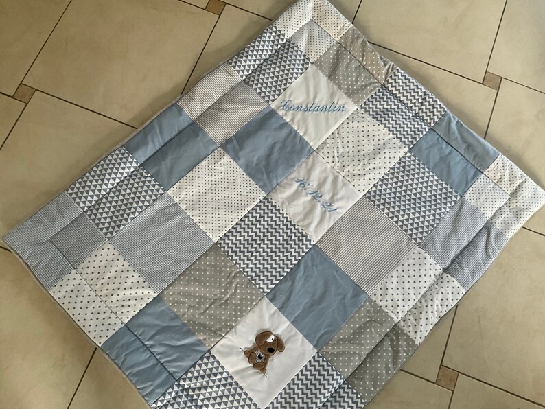 Peut inclure: Une courtepointe b&eacute;b&eacute; en patchwork bleu et gris avec un chien cousu et le nom "Constantin" et la date "16.12.2021".