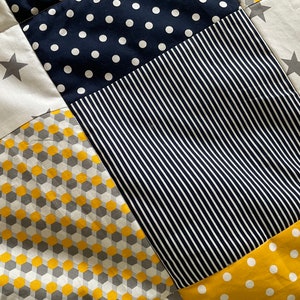 May include: A patchwork quilt featuring a range of patterns, including stars, polka dots, stripes, and geometric shapes. The quilt is made in shades of yellow, grey, and navy blue.