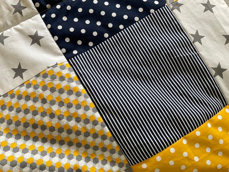 May include: A patchwork quilt featuring a range of patterns, including stars, polka dots, stripes, and geometric shapes. The quilt is made in shades of yellow, grey, and navy blue.