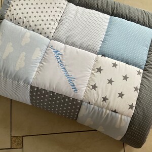 May include: A blue and gray patchwork baby quilt with a white square in the center that says "Maximilian" in blue embroidery.