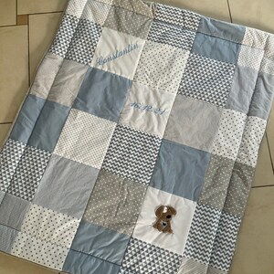 Peut inclure: Une courtepointe b&eacute;b&eacute; en patchwork bleu et blanc avec une bordure &agrave; motifs gris et blanc. La courtepointe a un petit chien brod&eacute; au centre et le texte "Constantin 16.12.21" brod&eacute; en haut au centre.