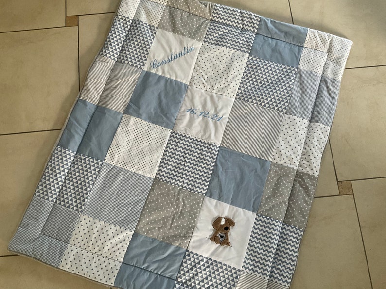 Peut inclure: Une courtepointe b&eacute;b&eacute; en patchwork bleu et blanc avec une bordure &agrave; motifs gris et blanc. La courtepointe a un petit chien brod&eacute; au centre et le texte "Constantin 16.12.21" brod&eacute; en haut au centre.