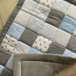 May include: A gray and blue patchwork baby playmat with a personalized name, Maximilian, embroidered in the center. The playmat has a soft, gray backing.
