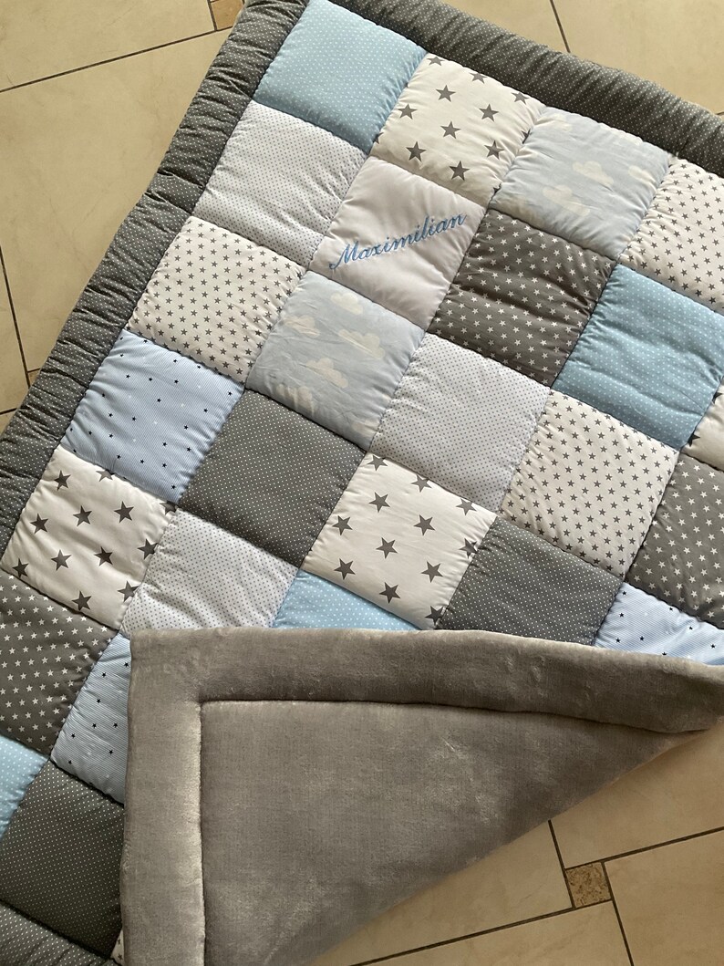 May include: A gray and blue patchwork baby playmat with a personalized name, Maximilian, embroidered in the center. The playmat has a soft, gray backing.