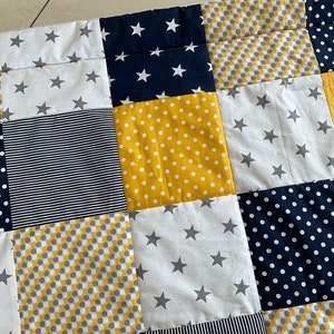 May include: A patchwork quilt featuring a range of patterns in yellow, white, grey, and navy blue. The patterns include polka dots, stripes, stars, and geometric shapes.