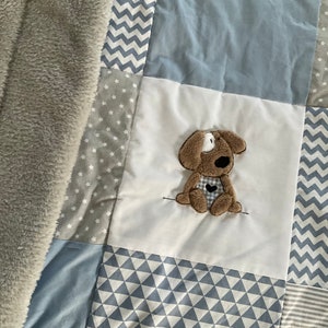 Peut inclure: Une couverture b&eacute;b&eacute; en patchwork de couleur gris, blanc et bleu. La couverture pr&eacute;sente une vari&eacute;t&eacute; de motifs, notamment des &eacute;toiles, des zigzags et des triangles. Une applique en forme de chien en peluche marron est centr&eacute;e sur la couverture.