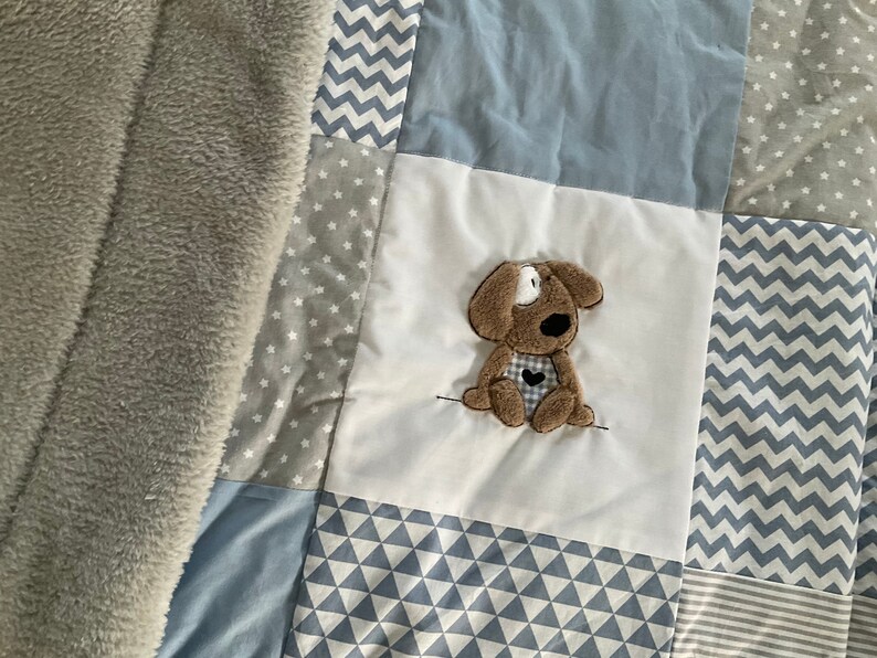 Peut inclure: Une couverture b&eacute;b&eacute; en patchwork de couleur gris, blanc et bleu. La couverture pr&eacute;sente une vari&eacute;t&eacute; de motifs, notamment des &eacute;toiles, des zigzags et des triangles. Une applique en forme de chien en peluche marron est centr&eacute;e sur la couverture.