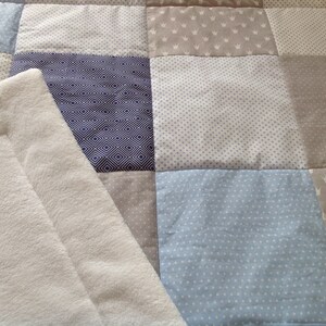 Könnte beinhalten: Eine weiße, plüschige Babydecke mit Patchwork-Muster. Die Decke besteht aus verschiedenen Stoffen mit verschiedenen Mustern, darunter blaue und weiße Punkte, grau mit weißen Kronen und blaue und weiße geometrische Muster.