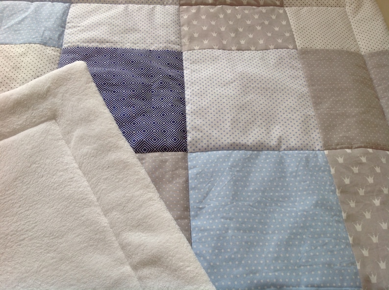 Pu&ograve; includere: Una morbida coperta per neonati di colore bianco con un design a patchwork. La coperta &egrave; realizzata con diversi tessuti con vari motivi, tra cui pois blu e bianchi, grigio con corone bianche e motivi geometrici blu e bianchi.