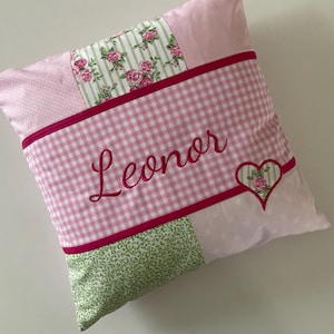 Könnte beinhalten: Ein quadratisches Patchwork-Kissen in Rosa- und Grüntönen. Das Kissen hat ein Vichy-Muster, Blumendrucke und den gestickten Namen "Leonor" in Schreibschrift. Ein herzförmiges Applikation mit floralen Details sorgt für eine dekorative Note.