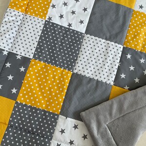 May include: A patchwork quilt in a grey, white and yellow colour scheme. The quilt is made of squares with different patterns, including stars, polka dots and small stars. The quilt is folded in half and has a grey fleece backing.