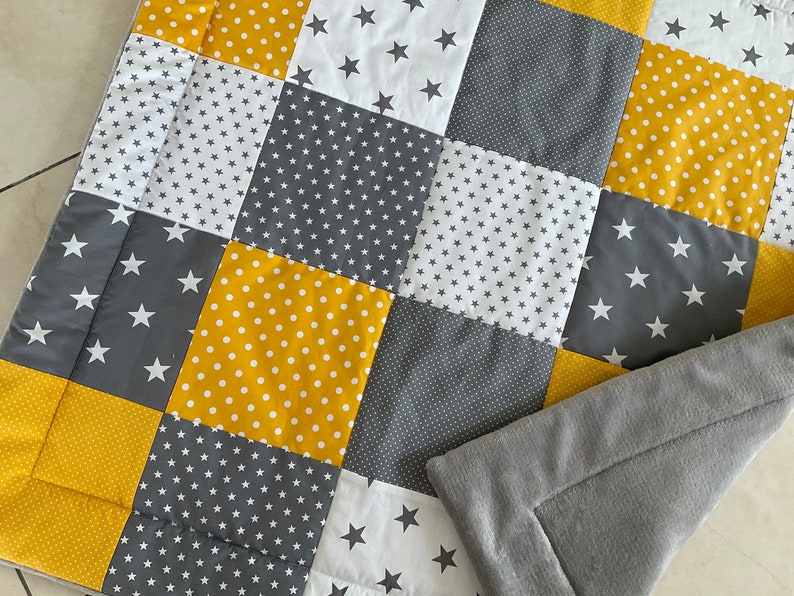 May include: A patchwork quilt in a grey, white and yellow colour scheme. The quilt is made of squares with different patterns, including stars, polka dots and small stars. The quilt is folded in half and has a grey fleece backing.