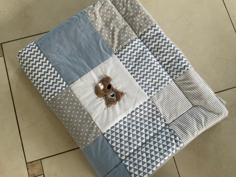 Peut inclure: Une couverture b&eacute;b&eacute; en patchwork bleu et gris avec un petit chien brun brod&eacute; au centre.