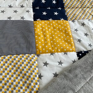 May include: A patchwork quilt featuring a range of patterns in yellow, white, grey, and navy blue. The quilt is made up of squares with different designs, including stars, polka dots, stripes, and geometric shapes. A grey blanket is partially covering the quilt.