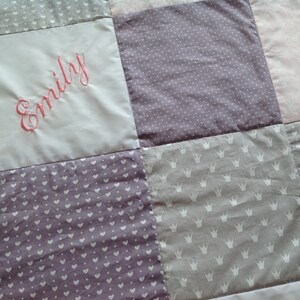 Pu&ograve; includere: Una trapunta patchwork nei toni del rosa, lavanda e grigio. La trapunta presenta quadrati con vari motivi, tra cui pois, cuori e corone. Il nome "Emily" &egrave; ricamato con filo rosa su un quadrato grigio chiaro.