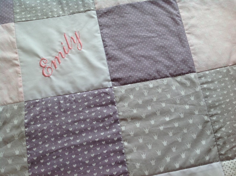 Pu&ograve; includere: Una trapunta patchwork nei toni del rosa, lavanda e grigio. La trapunta presenta quadrati con vari motivi, tra cui pois, cuori e corone. Il nome "Emily" &egrave; ricamato con filo rosa su un quadrato grigio chiaro.