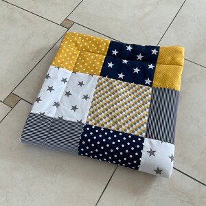 May include: A patchwork baby playmat featuring a variety of patterns in yellow, navy blue, white and grey. The patterns include stars, polka dots, stripes and geometric designs.