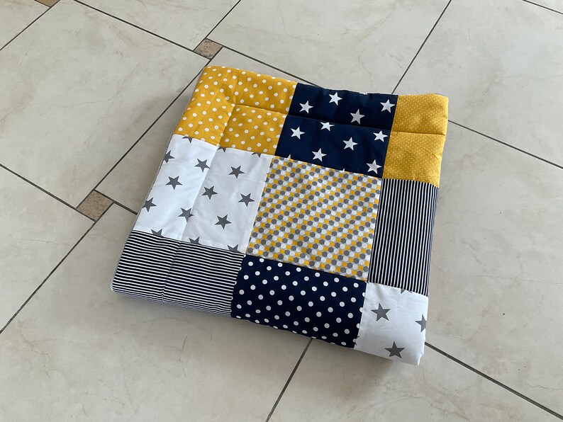 May include: A patchwork baby playmat featuring a variety of patterns in yellow, navy blue, white and grey. The patterns include stars, polka dots, stripes and geometric designs.