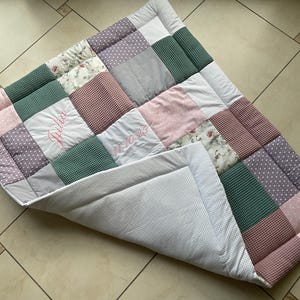 Könnte beinhalten: Ein Patchwork-Quilt mit verschiedenen Stoffen in Rosa, Grün, Grau und Weiß. Der Quilt hat Quadrate mit floralen, gepunkteten und strukturierten Mustern. Der Name "Julia" ist in Rosa auf einem Quadrat gestickt, und das Datum "14-10-23" ist auf einem anderen gestickt.