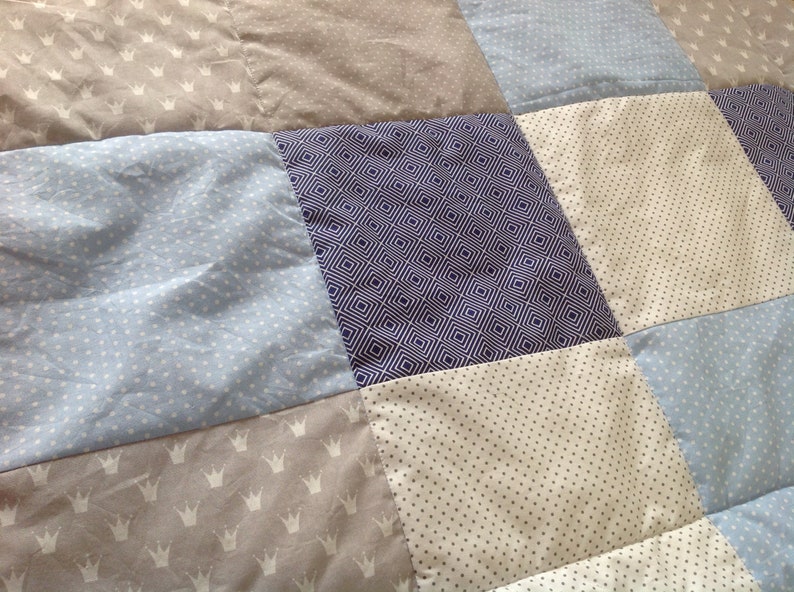 Pu&ograve; includere: Una trapunta patchwork con una variet&agrave; di motivi, tra cui corone, pois e disegni geometrici. La trapunta &egrave; realizzata in tonalit&agrave; di blu, grigio e bianco.
