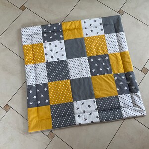 May include: A patchwork playmat in a yellow, grey and white colour scheme. The mat is made of squares with different patterns, including stars, polka dots and solid colours.