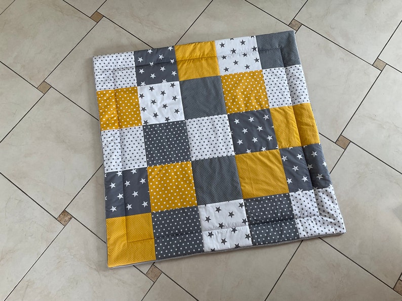 May include: A patchwork playmat in a yellow, grey and white colour scheme. The mat is made of squares with different patterns, including stars, polka dots and solid colours.