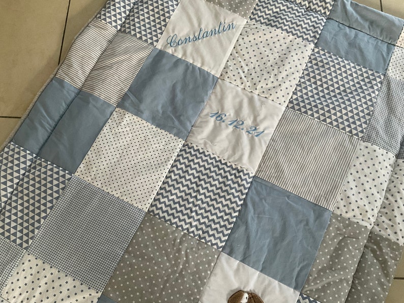 Peut inclure: Une courtepointe b&eacute;b&eacute; en patchwork bleu et blanc avec le nom "Constantin" et la date "16.12.21" brod&eacute;s dessus.