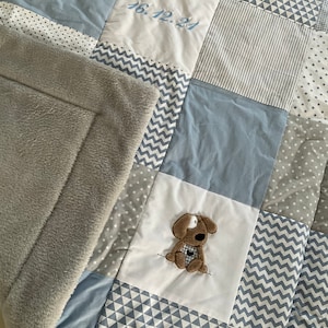Peut inclure: Une couverture b&eacute;b&eacute; en patchwork gris et bleu avec une applique de chien cousue et la date "16.12.21" brod&eacute;e sur l'un des carr&eacute;s.