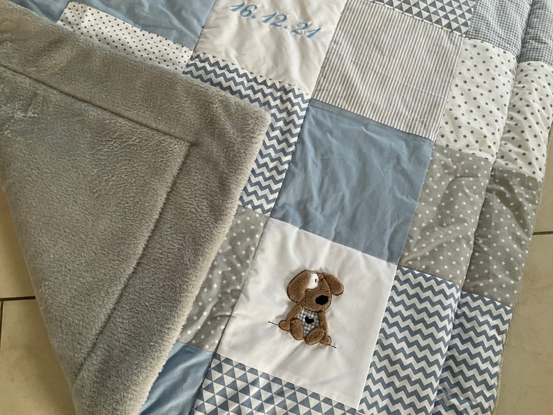 Peut inclure: Une couverture b&eacute;b&eacute; en patchwork gris et bleu avec une applique de chien cousue et la date "16.12.21" brod&eacute;e sur l'un des carr&eacute;s.