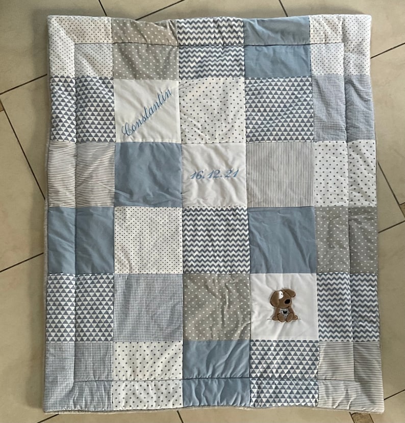 Peut inclure: Une courtepointe b&eacute;b&eacute; patchwork bleu et gris avec un chien brod&eacute; blanc et le texte "Constantijn 16.12.24".