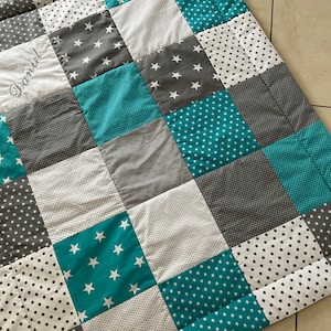 Könnte beinhalten: Ein Patchwork-Quilt mit Quadraten in Türkis, Grau, Weiß und Schwarz. Einige Quadrate haben weiße Sterne oder Punkte. Der Name "Daniel" ist auf einem weißen Quadrat gestickt. Ein handgefertigter, gemütlicher Artikel.