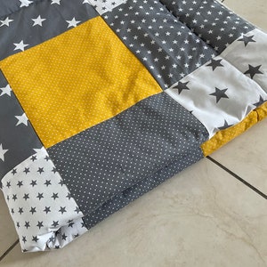 May include: A yellow, grey, and white patchwork quilt with a star pattern. The quilt is folded in half and is lying on a light grey surface.