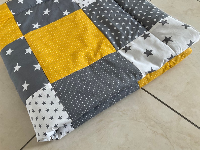 May include: A yellow, grey, and white patchwork quilt with a star pattern. The quilt is folded in half and is lying on a light grey surface.