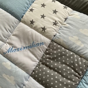 May include: A baby quilt with a patchwork design featuring different patterns, including stars, clouds, and polka dots. The quilt is personalized with the name "Maximilian" embroidered in blue thread.