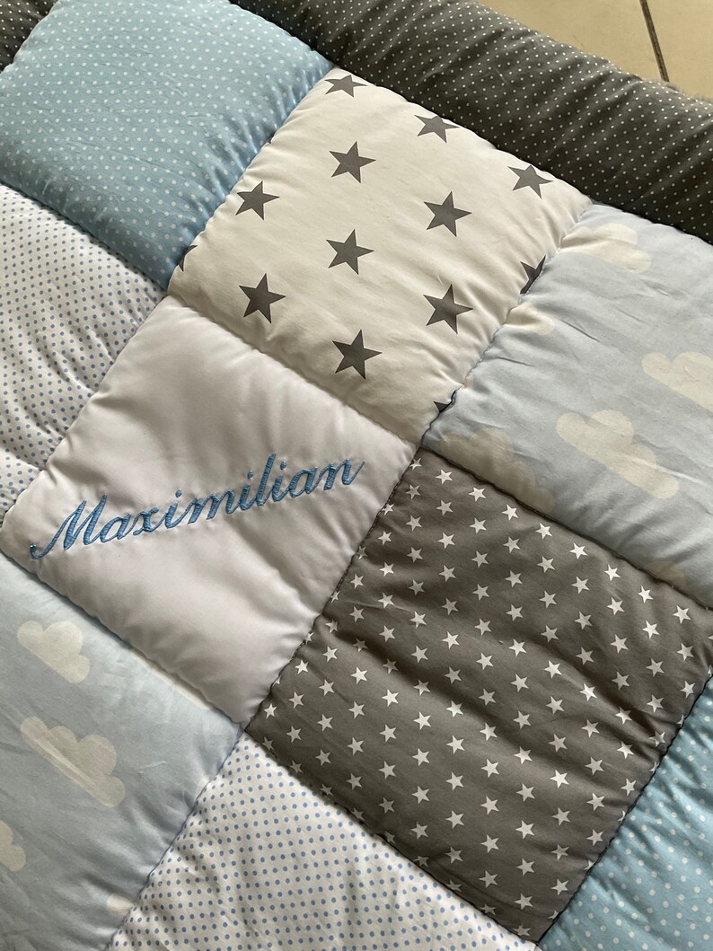 May include: A baby quilt with a patchwork design featuring different patterns, including stars, clouds, and polka dots. The quilt is personalized with the name "Maximilian" embroidered in blue thread.