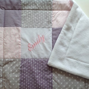 Pu&ograve; includere: Una coperta patchwork per bambini nei toni del rosa, grigio e viola. La coperta presenta un quadrato bianco con il nome "Emily" ricamato in rosa. Il retro &egrave; in morbido tessuto bianco.