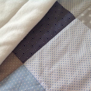 Pu&ograve; includere: Un quilt patchwork con un motivo bianco, beige, blu scuro e blu chiaro. Il quilt &egrave; realizzato con diversi tessuti con vari motivi, tra cui pois, quadrati e corone.