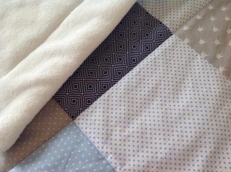 Pu&ograve; includere: Un quilt patchwork con un motivo bianco, beige, blu scuro e blu chiaro. Il quilt &egrave; realizzato con diversi tessuti con vari motivi, tra cui pois, quadrati e corone.