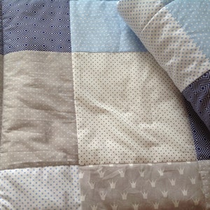 Pu&ograve; includere: Un quilt patchwork con una variet&agrave; di motivi in ​​sfumature di blu, grigio e bianco. Il quilt &egrave; fatto di quadrati con diversi motivi, tra cui pois, diamanti e corone.