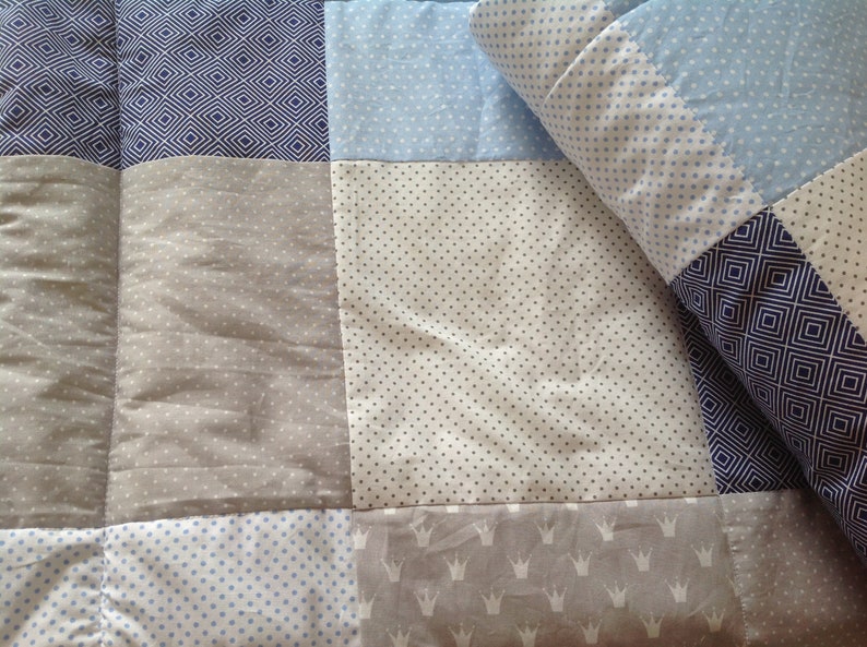 Pu&ograve; includere: Un quilt patchwork con una variet&agrave; di motivi in ​​sfumature di blu, grigio e bianco. Il quilt &egrave; fatto di quadrati con diversi motivi, tra cui pois, diamanti e corone.