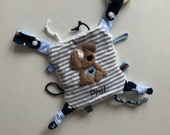 May include: A baby sensory toy with a central square featuring a brown dog applique on a striped background. The toy has various patterned tabs in blue and white, with the name "Phil" embroidered on it.