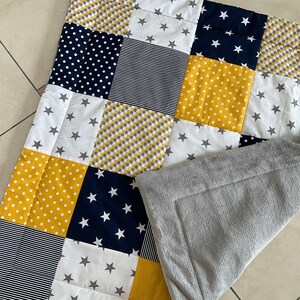 May include: A patchwork baby blanket with a grey fleece backing. The blanket is made of various fabrics in yellow, white, navy blue, and grey with different patterns including polka dots, stripes, and stars.